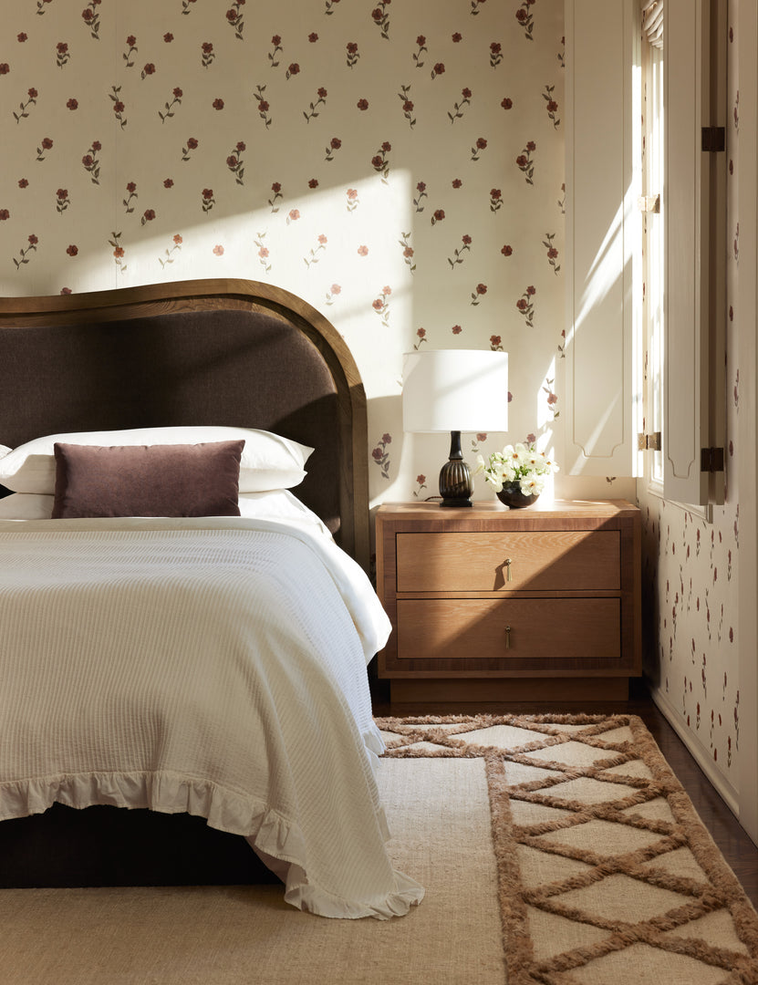 #color::red | A wavy headboard bed frame and wooden nightstand sit in front of a wall covered with the Bamonte Textile String Floral Wallpaper by Jake Arnold