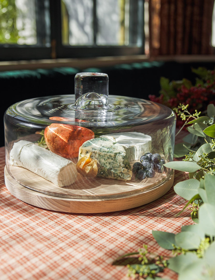 #style::pastry-dome | The Lotta cheese and pastries glass dome with ash wood base by LSA International sits in a dining room atop a red plaid tablecloth next to an olive garland