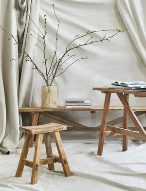 The Arlene craftsman-style antiqued teak wood bench, along with the matching Arlene stool and console table are displayed against a wall draped in light tan fabric.