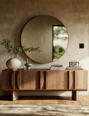 A large round mirror hangs above the Remwald sculptural oak wood low profile media console.