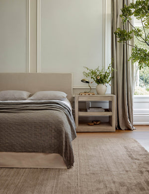 The Chiltern sand dune rug lays in a bright bedroom under a natural linen framed bed and a light wooden night stand
