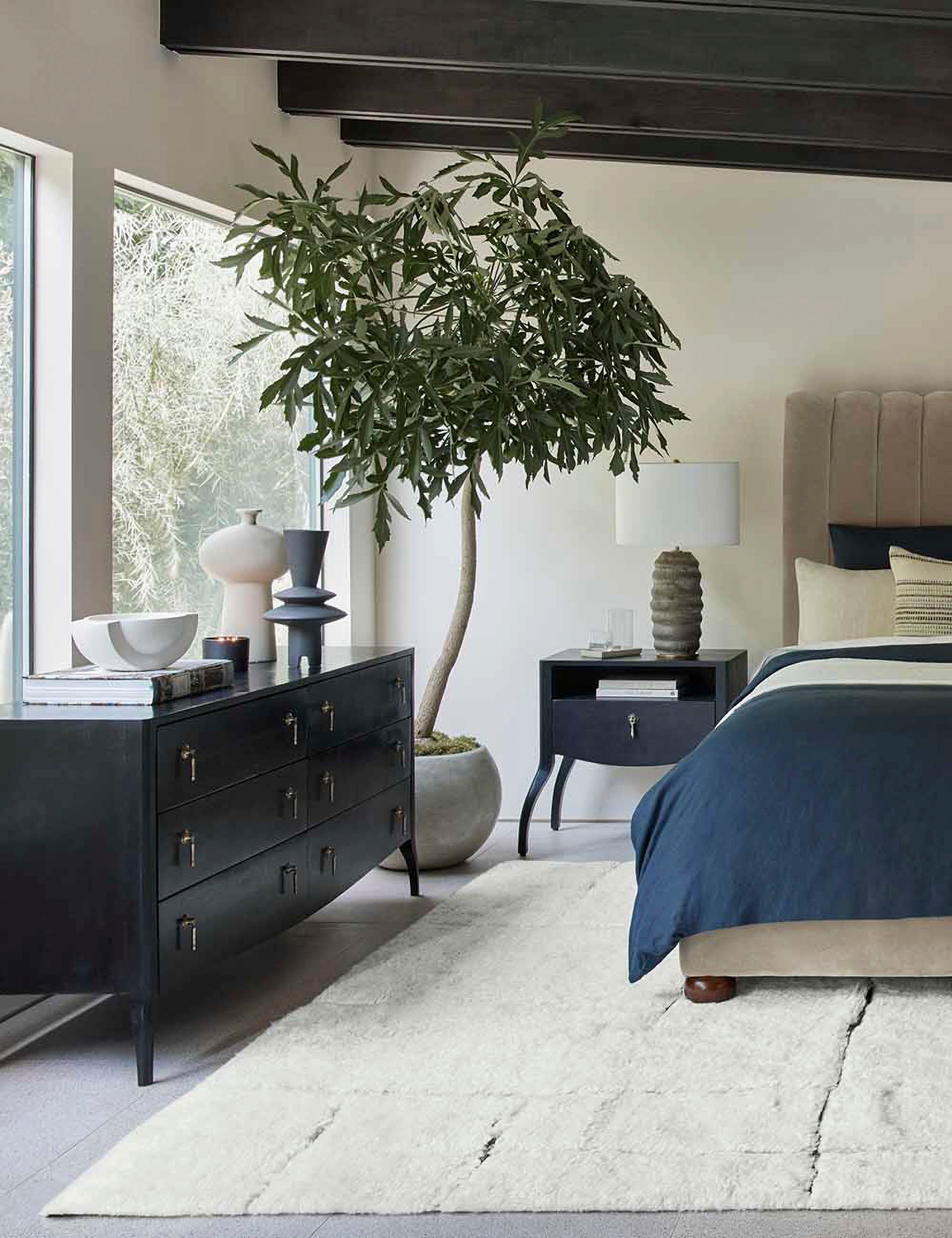 | The Anabella black wood dresser with silver drawer pulls sits in a bedroom underneath two sculptural vases with a gray velvet bed and anabella nightstand to the right of it.