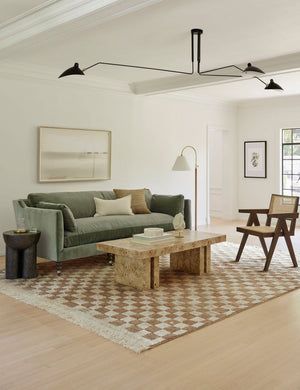 The Brisa rectangular burl wood coffee table with four legs sits in a modern living room with a moss green sofa, a tan and ivory checked rug, and a brown wood and cane accent chair under a large modern black chandelier.