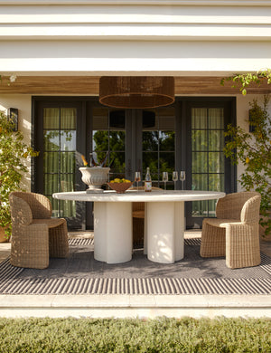 Rodrigo sculptural oval outdoor dining table styled with two wicker outdoor dining chairs in a covered outdoor space.
