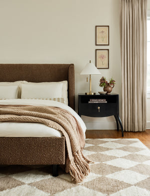 The Anabella black wood nightstand sits to the right of an ochre boucle framed bed in front of a carpet with a diamond pattern