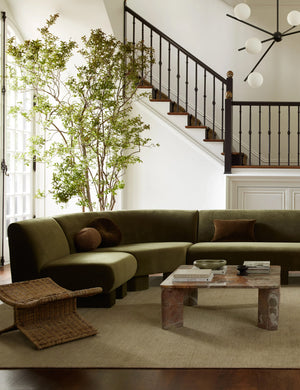 The Heritage moss gray rug lays in a living room under a olive green velvet sectional, a woven ottoman, and a stone coffee table