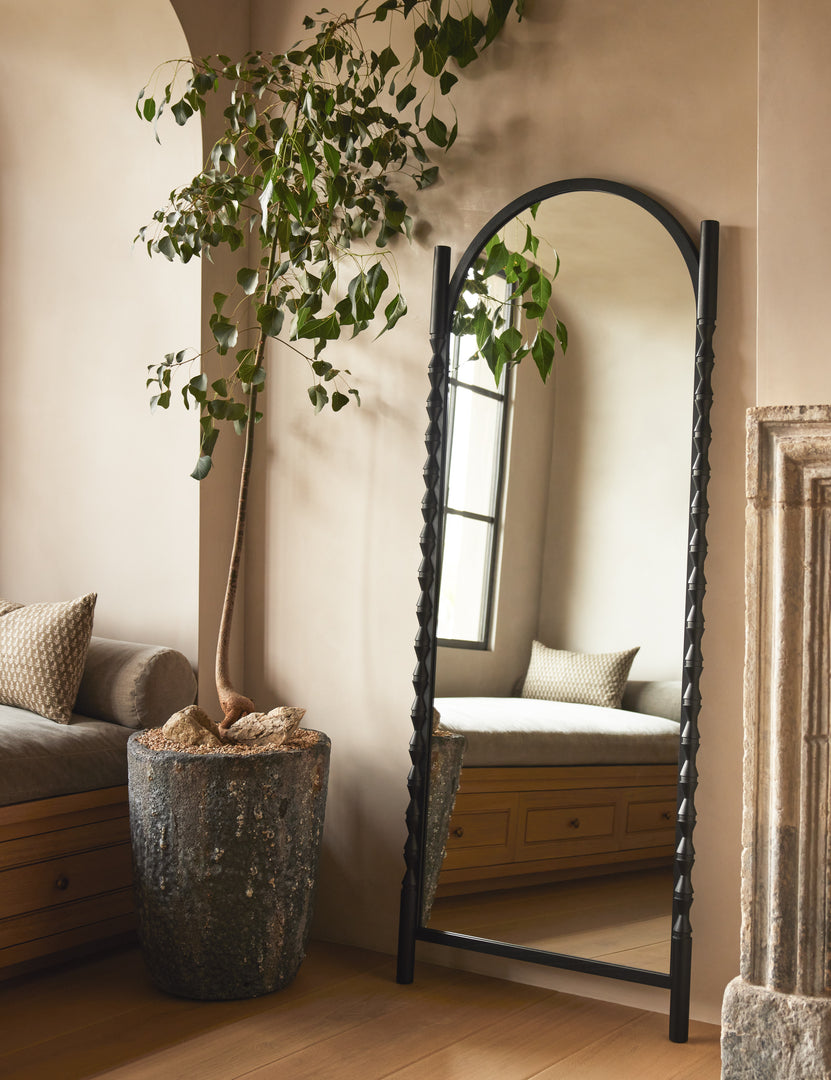 #color::black | Topia arched carved wood frame floor mirror by Ginny Macdonald in black leaning against a wall next to a decorative plant.