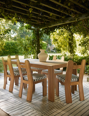 Six Abbot solid teak sculptural outdoor dining chair by Sarah Sherman Samuel surround a teak outdoor dining table under a pergola.