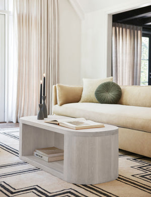 The Luna white-washed oak oval coffee table sits in a minimal living room atop a geometric rug with wooden candles and a book sitting on its surface.