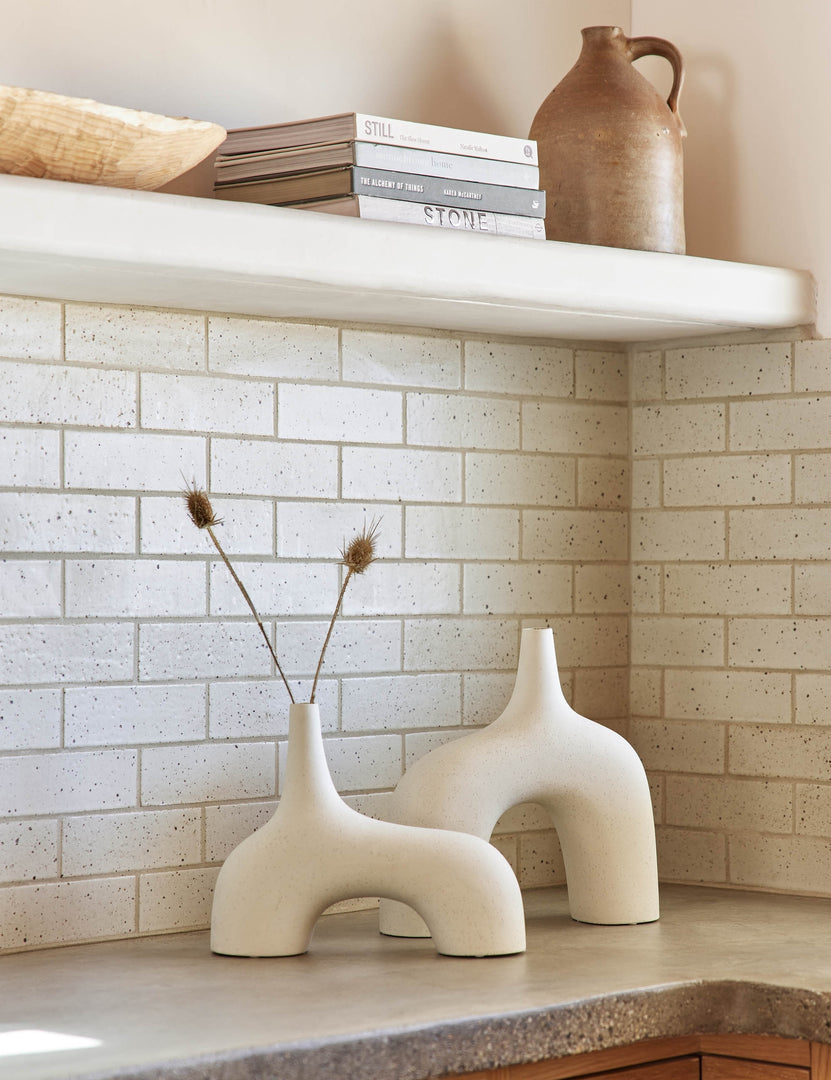 #color::white #size::small  #size::large | The Leonor sculptural arched matte white ceramic Vase in its small and large size sitting atop a granite countertop in a kitchen with a subway tile backsplash, a stack of books, and a wooden centerpiece bowl