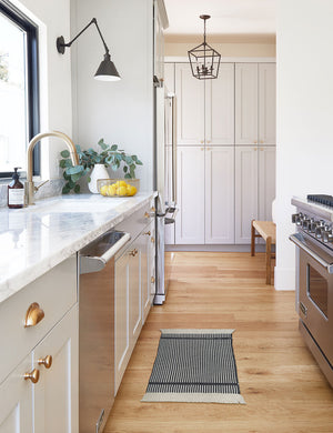 The Ines handwoven black-and-white striped mat lays on a wooden floor in a kitchen with white cabinetry and marble countertops