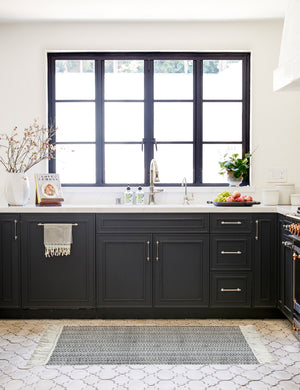 The Saskia handwoven high contrast black-and-white patterned mat with thick fringe lays on a kitchen floor surrounded by marble counter tops and black accented cabinets