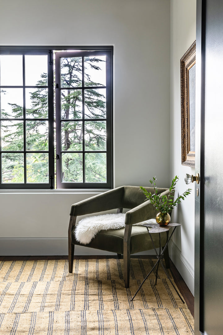 | The Lyssa olive velvet accent chair sits in the corner of a room with a striped rug and a white sheepskin blanket thrown atop it.