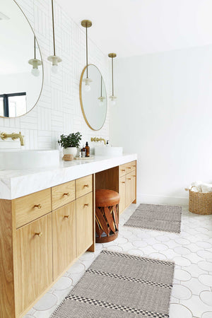Two Masha gray and black geometric machine washable mats lay in a bathroom with circular golden mirrors and marble countertops