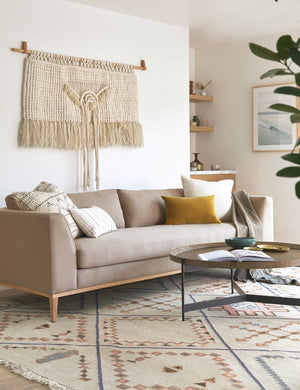 The Charleston pebble gray linen sofa sits under a woven wall hanging atop of an ivory rug with a diamond patterned design