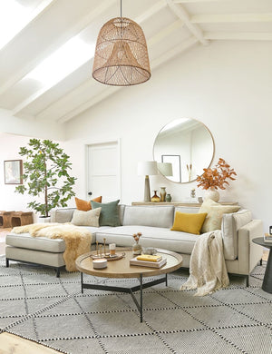 The Becca coffee table with rimmed oak top and iron tripod-style base sits in a living room atop a black and white patterned rug in front of a linen sofa underneath a natural jute chandelier
