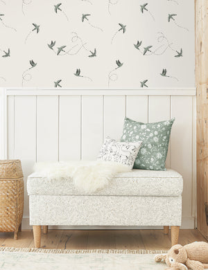 The Sparrow blue and white wallpaper by Rylee and Cru is in a bedroom above a white wooden paneled wall with a floral patterned bench and patterned throw pillows.