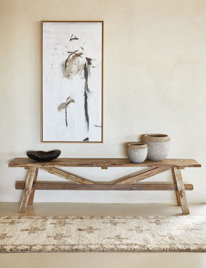 An Acoma cream and tan plus-sign patterned Moroccan runner rug with diamond border lays in front of a light wood craftsman-style bench with an abstract black, gray and white painting hung above.