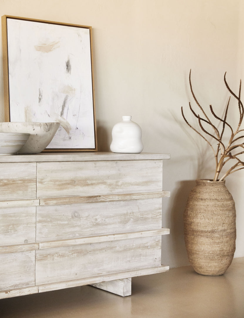 | The Corliss 6-drawer white-washed wooden dresser has a white abstract painting leaning on top of it and decorative white bowls and vases.