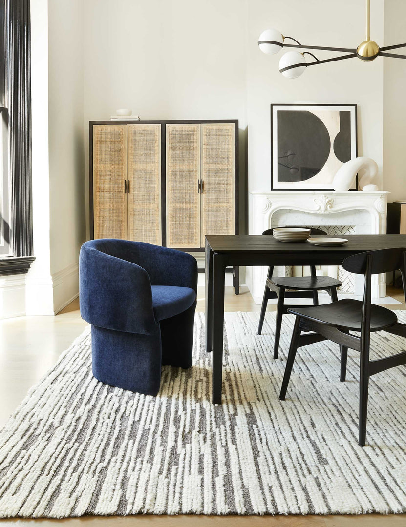 | The Adeline black and brass retro chandelier sits in a dining room with a black wooden dining table, black wooden dining chairs, and a navy velvet accent chair