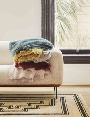 The Aimee mohair wool throw in mustard, blue, blush, warm gray, and merlot are stacked atop each other on a gray sofa in a living room with a geometric rug