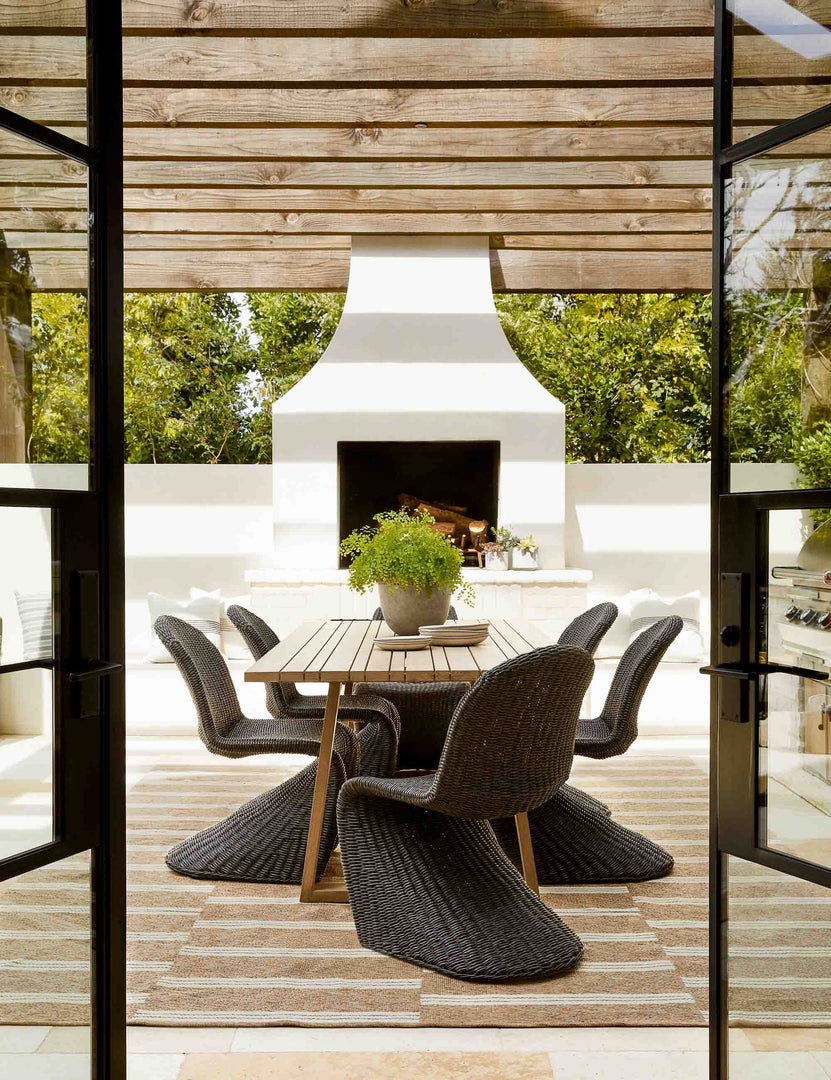 #color::Vintage-Coal  | The Manila wicker weave black indoor and outdoor dining chair sits in an outdoor dining space underneath a wood paneled covering with a large white fireplace in the background.