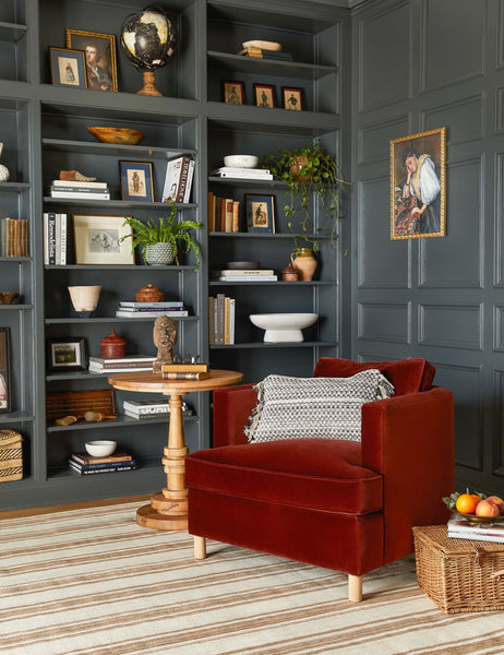 #color::paprika | The Belmont paprika red velvet accent chair sits atop a striped rug in front of a gray wall with a built-in shelf and decor