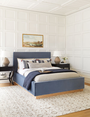  The Lockwood blue velvet-upholstered bed with a white oak base sits atop a patterned hand-woven rug against a white, accented wall.