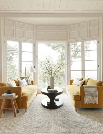 #showall::39490333343843 | The Elulia woven two-tone taupe throw with fringed ends lays atop a yellow velvet sofa in a living room with an oval coffee table, white accented walls, and floor to ceiling french doors and windows