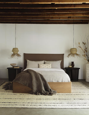 The Nia mushroom brown linen bed sits atop a plush rug in between two jute pendant lights under a wooden beamed ceiling