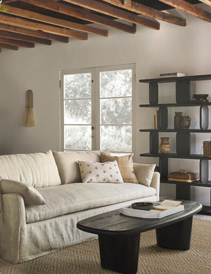 The Rica taupe square throw pillow with blue, white, and black woven arrow-like designs sits on a linen sofa next to a black oval coffee table