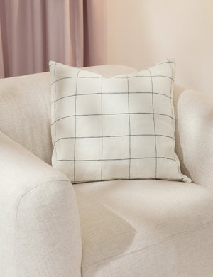 The lucian white and black square pillow sits atop an ivory accent chair in a studio room with purple curtains