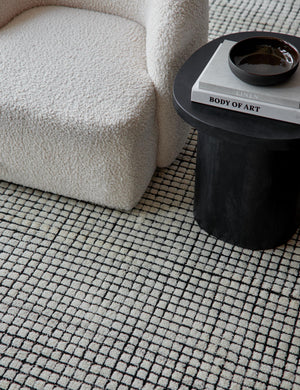 The Uma black and natural checkered grid pattern rug lays under a cream boucle accent chair and a round black wooden side table