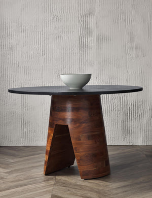 The Adler acadia wooden round dining table sits in a studio with a white bowl atop chevron floors in front of a textured wall