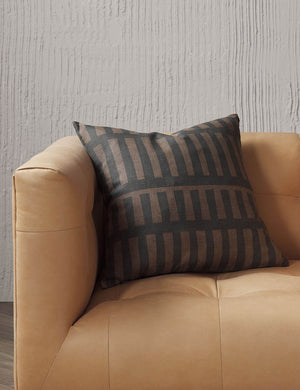 Kellan square throw pillow sits on a light brown leather sofa in a studio room with textured walls