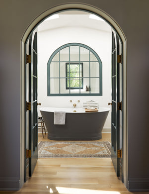 The Kehoe desert palette geometric floor rug sits in a bathroom in front of a black bathtub behind a black-framed arched entrance.