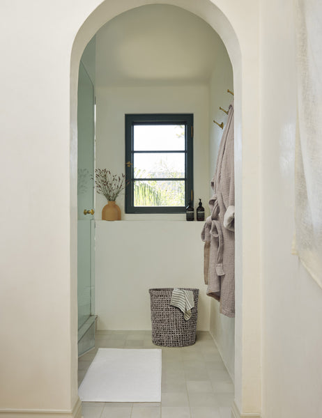 #color::white | The Cloud loom sustainable bath mat by coyuchi lays on a white tiled floor in a bathroom with an arched entryway