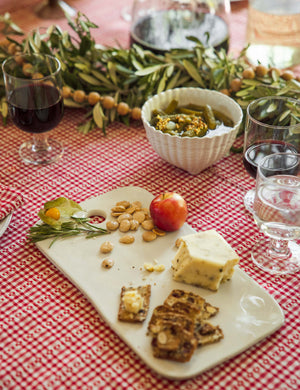 The Aparte white footed bowl with shell inspired design by Costa Nova sits on a red plaid tablecloth in a dining room with a marble cheese board and clear wine glasses