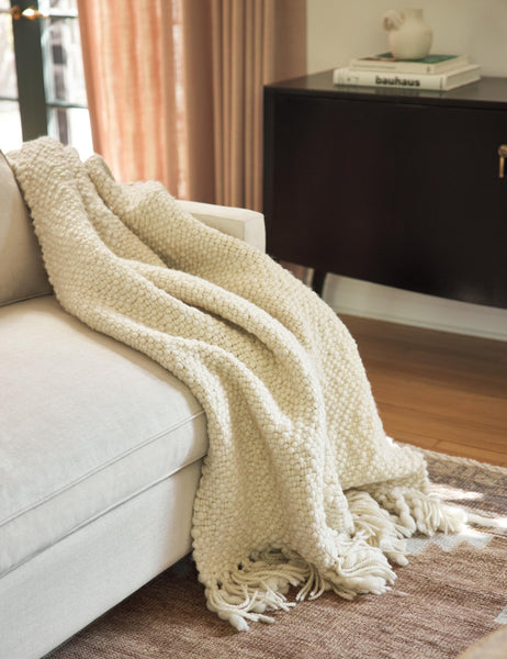 #color::ivory | The Olema ivory handwoven throw with fringed ends lays in a living room atop a natural linen sofa and a brown area rug with a black sideboard in the background