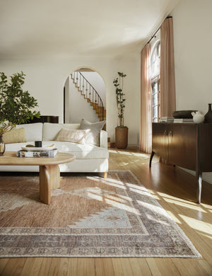 The Kehoe desert palette geometric floor rug sits in a living room with a white linen sofa, natural wooden coffee table with sculptural legs, and a wooden anabella console table.