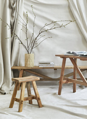 The Arlene craftsman-style antiqued teak wood bench, along with the matching Arlene stool and console table are displayed against a wall draped in light tan fabric.