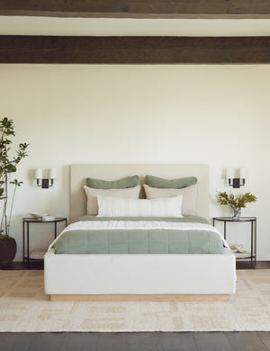  The Lockwood natural velvet-upholstered bed with a white oak base sits in a neutral bedroom with a plush natural rug and wooden ceiling beams. 