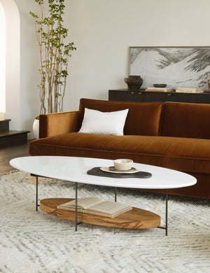 The Thomas Bina oval coffee table with white laquered top, oak shelf and steel frame sits in front of a dark rust velvet sofa and on top of a neutral area rug.