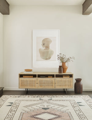 The Hannah natural wood media console with cane doors sits in an open space with a large portrait wall art mounted above it and a geometric woven rug below it.