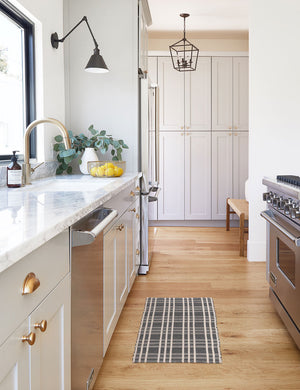 The Rika black and white cotton machine washable mat with checkerboard pattern in its runner size lays in a kitchen with marble countertops