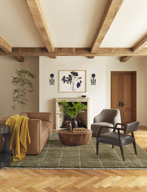 Khaki green Irregular Grid Rug by Sarah Sherman Samuel lays in a rustic living room under a tan velvet sofa and a round wooden coffee table