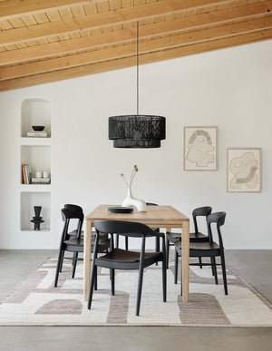 The Reese oak wood rectangular dining table sits in a dining room below a black jute chandelier surrounded by black dining chairs and accented by sculptural vases and wall art.