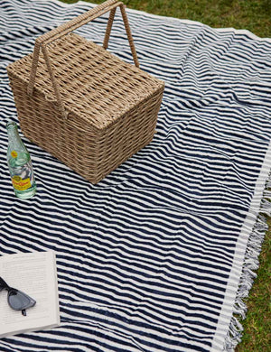 The Navy and white striped cotton beach blanket by business and pleasure co lays on the grass with a woven picnic basket