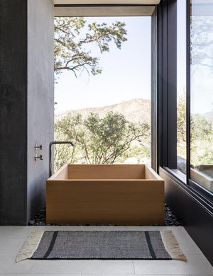 The Ines handwoven black-and-white striped mat lays on a gray tiled floor in a bathroom with a wooden square bathtub and floor to ceiling windows