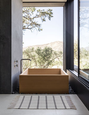 Elin mat lays on a tile floor in a bathroom with a rectangular wooden bath, black walls, and floor to ceiling windows
